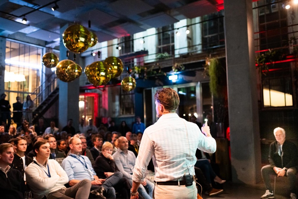 Spreker voor een zaal met publiek tijdens een AI-sessie in een moderne eventlocatie, met gouden decorbollen