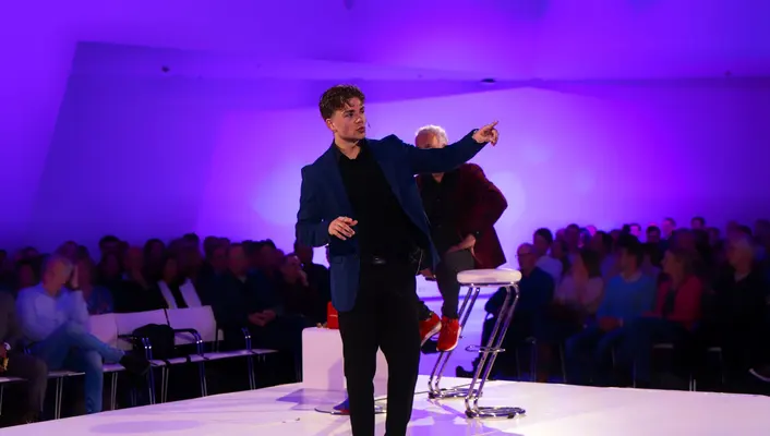Spreker wijst naar het publiek op een paars verlicht podium